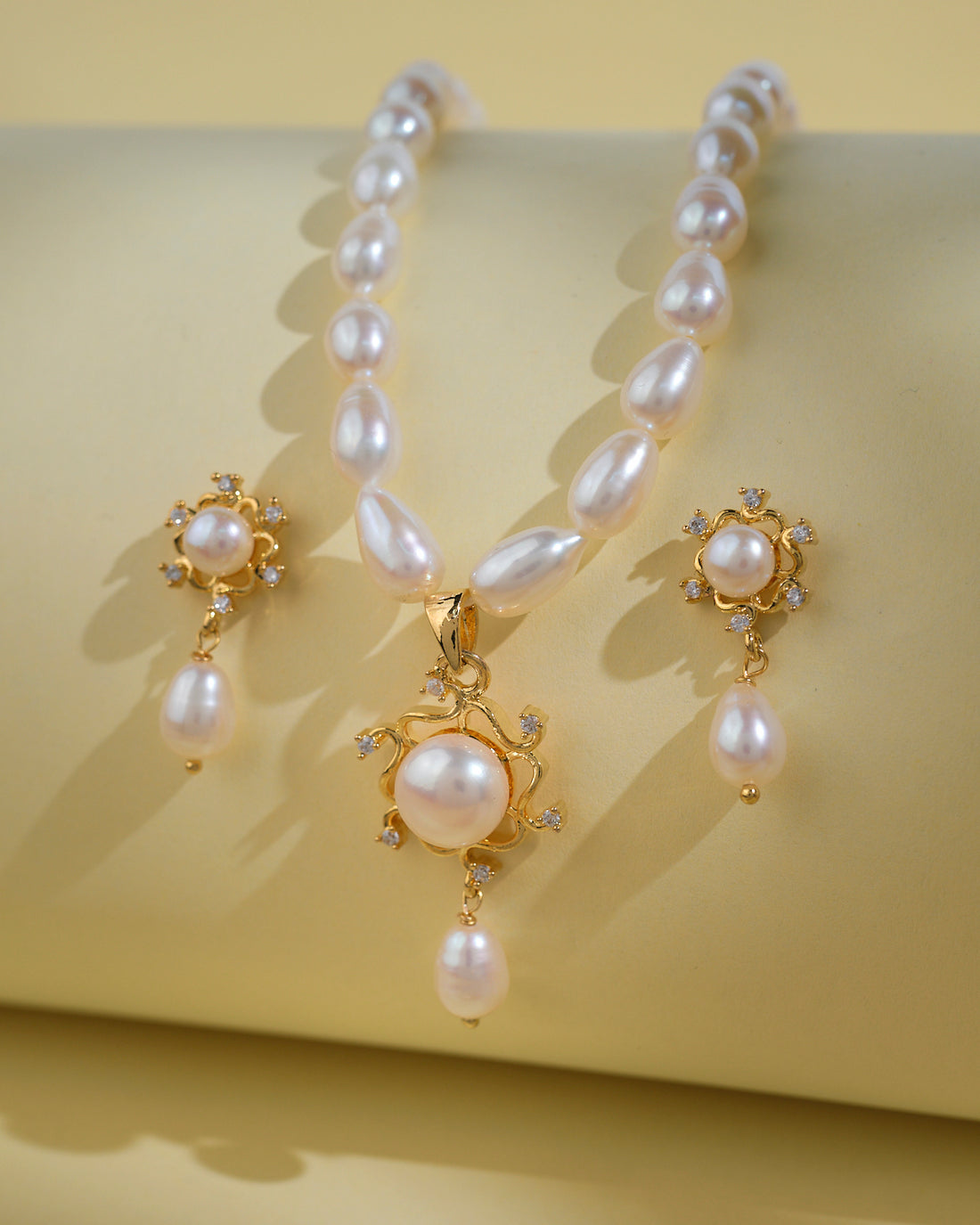 The Trendy Pearl Necklace Sets by Chandrani Pearls India feature elegant teardrop pearls and sparkling stones in a refined necklace and earrings set, perfect for the modern woman, shown on a pale yellow background.