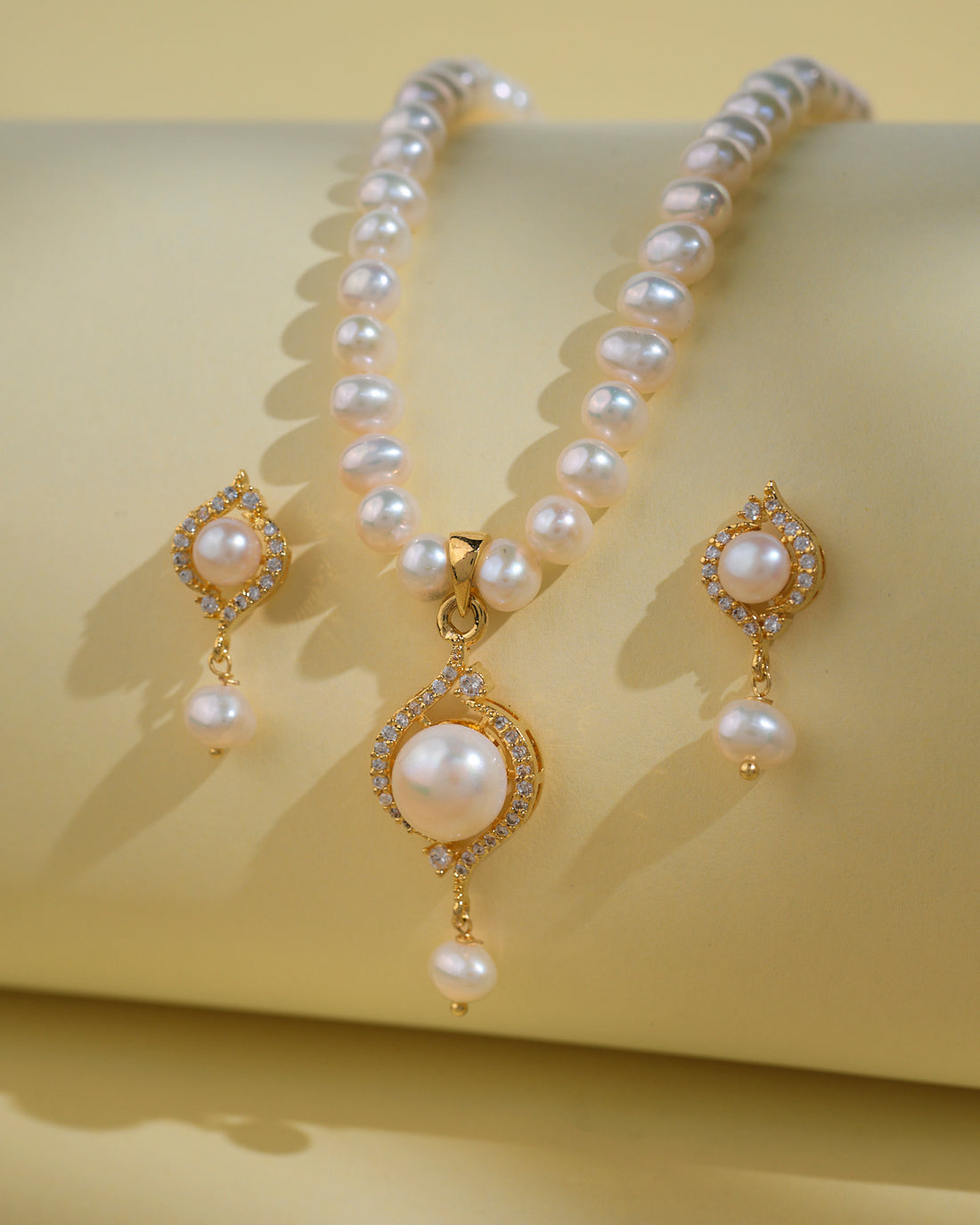 The Simple Pearl Necklace Sets by Chandrani Pearls India include a pearl necklace with gold and diamond accents and matching earrings, elegantly displayed on a pale yellow surface to enhance their natural beauty.