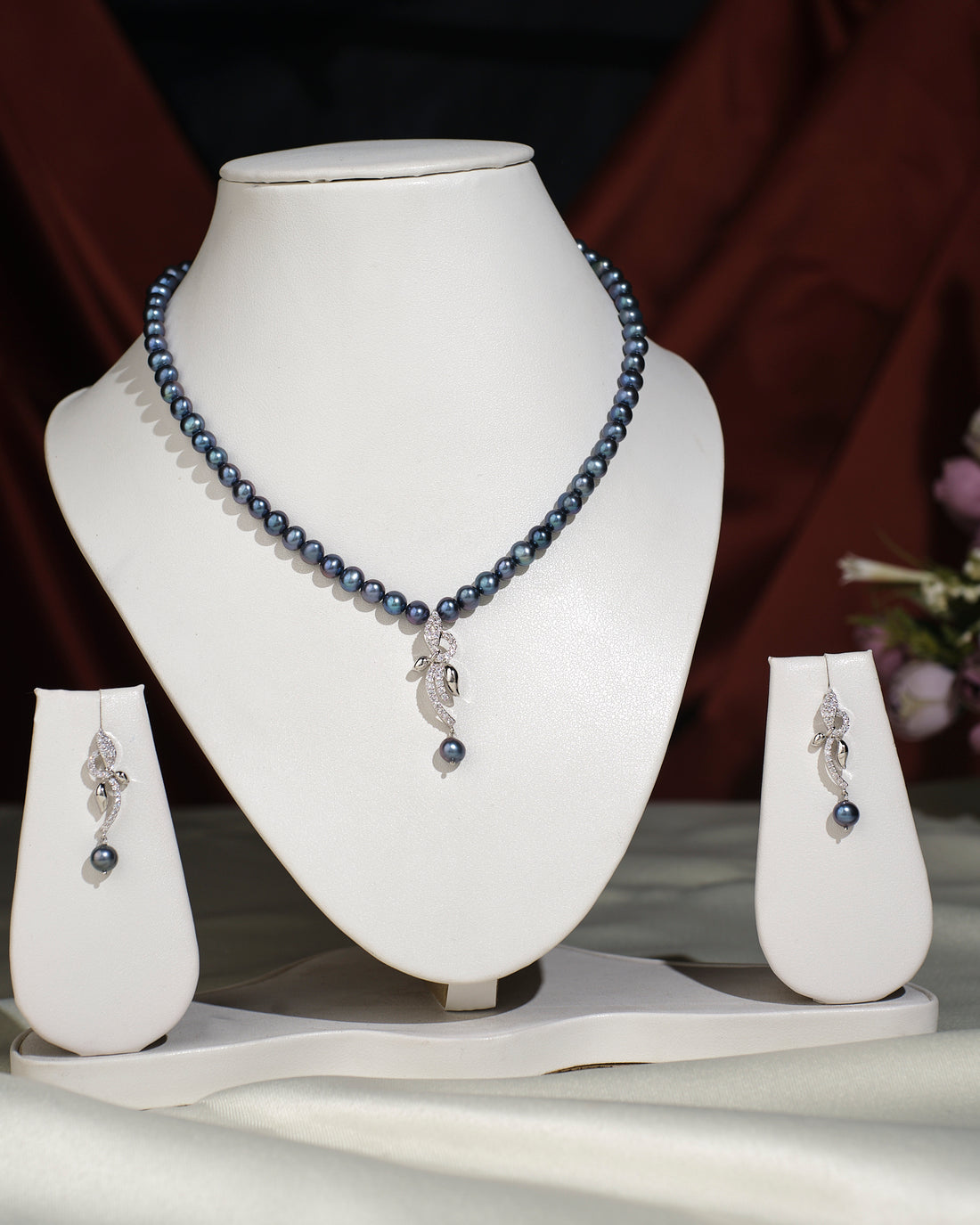 The Trendy Pearl Necklace Set by Chandrani Pearls India features a dark pearl necklace with a leaf pendant and matching earrings, elegantly displayed on stands against a fabric backdrop for a timeless look.