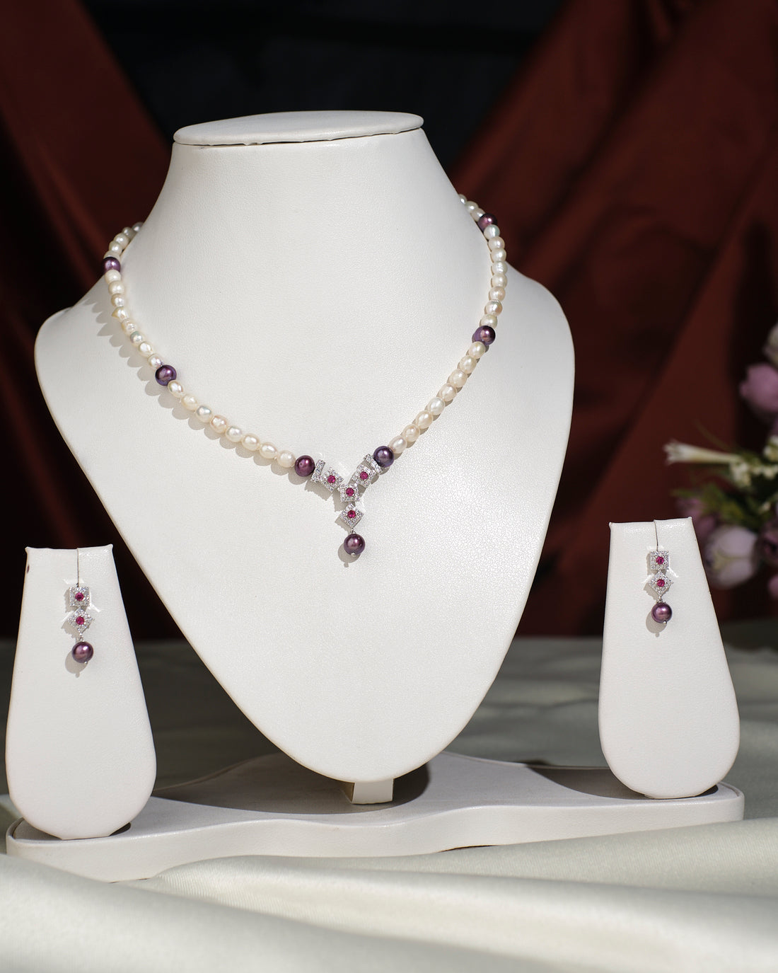 The Trendy Pearl Necklace Set by Chandrani Pearls India features pearls and amethyst beads with a matching pendant, shown on a white stand, alongside coordinating drop earrings displayed on earring stands.