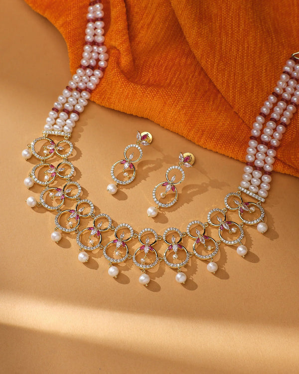 The Ravishing Real Pearl Bridal Jewelry Set by Chandrani Pearls features circular designs, pink accents, and pearl drops—ideal bridal jewelry. Displayed on a tan surface with an orange fabric backdrop.