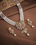 The Majestic Pearl & CZ Necklace Set by Chandrani Pearls features intricate designs, red and green gemstones, and matching earrings, sparkling brilliantly on brown embroidered fabric—perfect for any occasion.