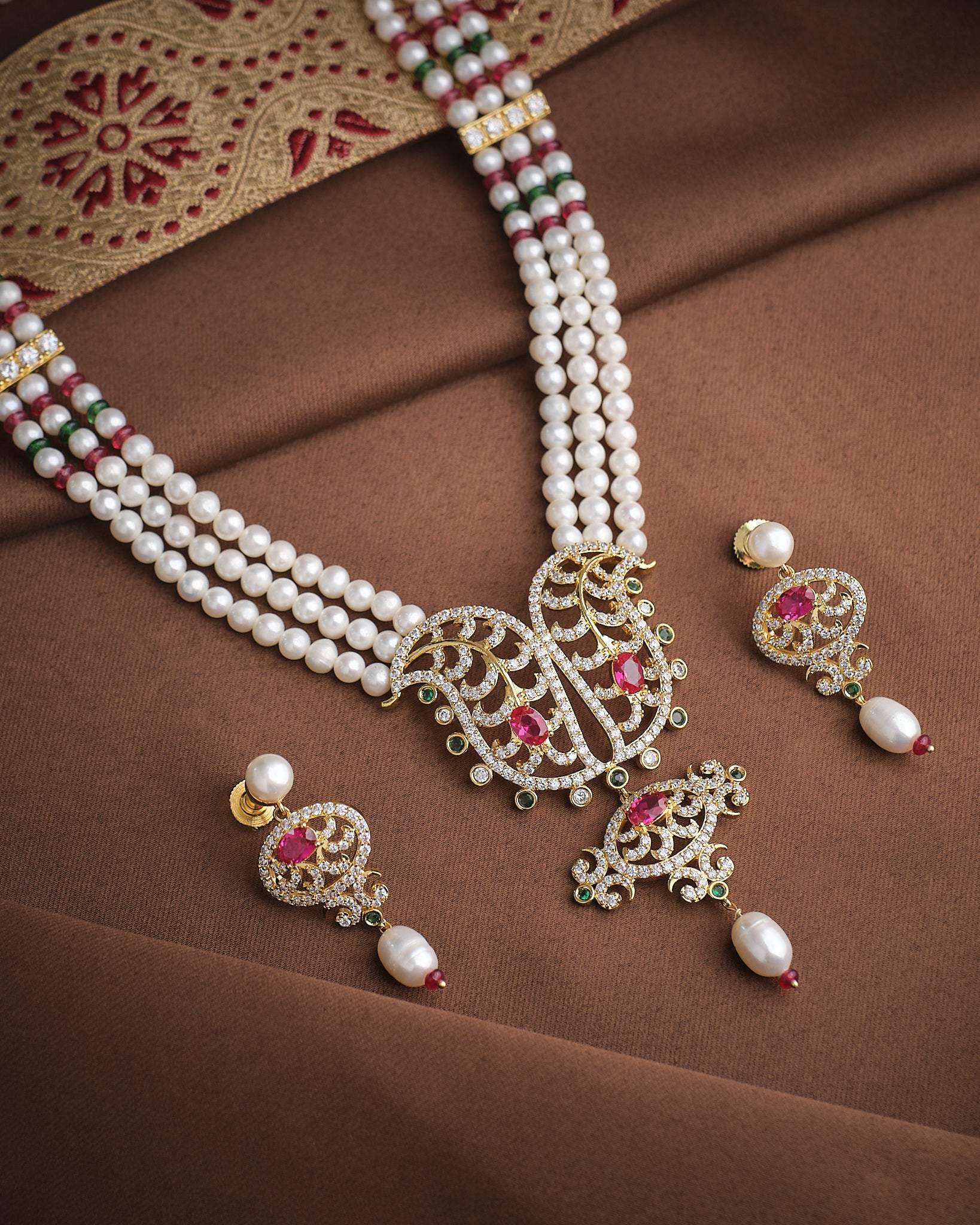 The Majestic Pearl & CZ Necklace Set by Chandrani Pearls features intricate designs, red and green gemstones, and matching earrings, sparkling brilliantly on brown embroidered fabric—perfect for any occasion.