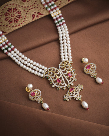 The Majestic Pearl & CZ Necklace Set by Chandrani Pearls features intricate designs, red and green gemstones, and matching earrings, sparkling brilliantly on brown embroidered fabric—perfect for any occasion.