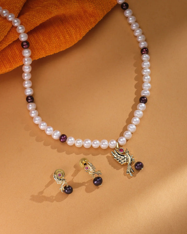 The Chandrani Pearls Classic Elegance Real Pearl Necklace Set, featuring gold and maroon accents, is displayed with matching earrings on a brown surface with orange fabric, highlighting the lustrous pearls.