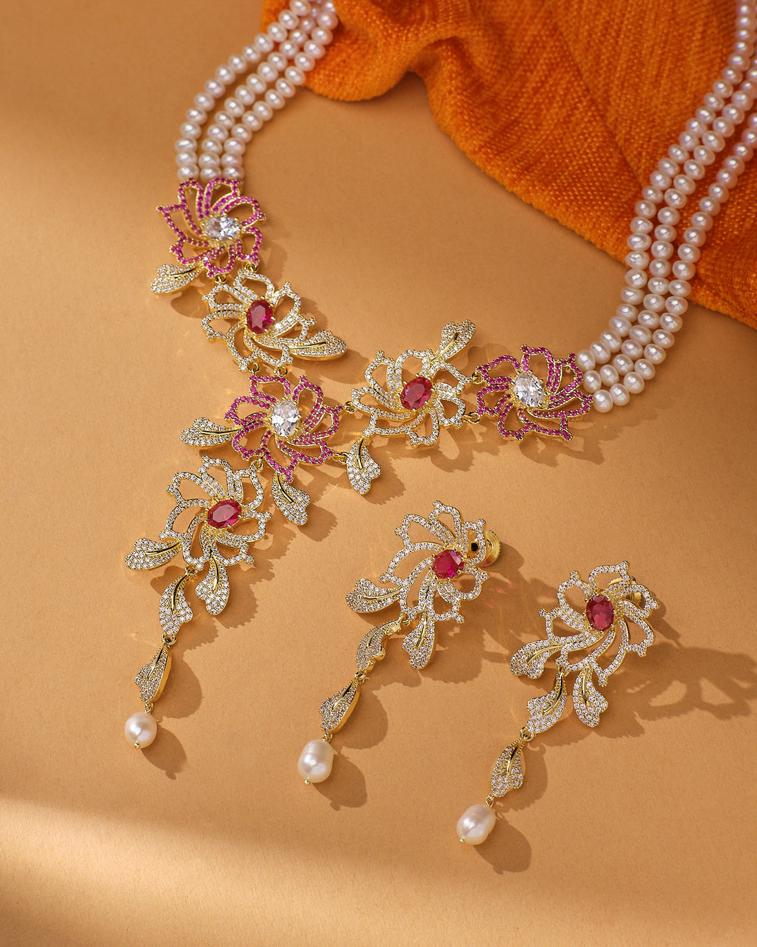 The Blushing Floral CZ Pearl Beauty Necklace Set by Chandrani Pearls features real pearls, gold and pink floral designs, red gemstones, and matching earrings on a beige backdrop with orange cloth—perfect for adding feminine charm to party evenings.
