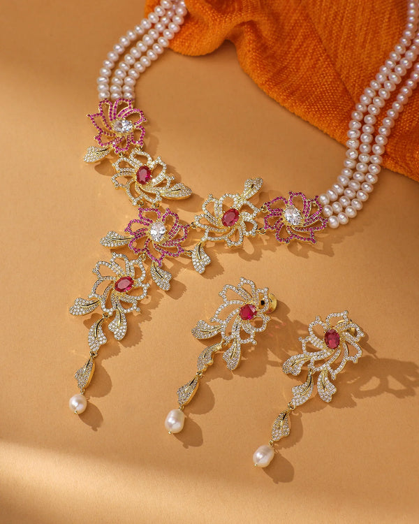 The Blushing Floral CZ Pearl Beauty Necklace Set by Chandrani Pearls features real pearls, gold and pink floral designs, red gemstones, and matching earrings on a beige backdrop with orange cloth—perfect for adding feminine charm to party evenings.
