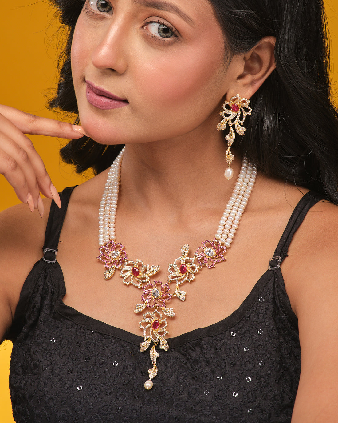 The Blushing Floral CZ Pearl Beauty Necklace Set by Chandrani Pearls features real pearls, gold and pink floral designs, red gemstones, and matching earrings on a beige backdrop with orange cloth—perfect for adding feminine charm to party evenings.