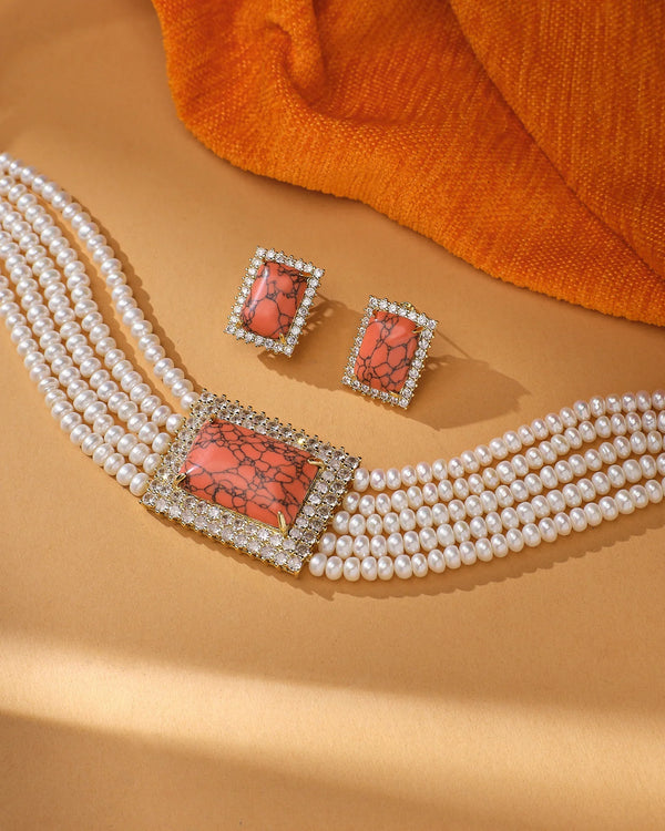 The Trendsetter Pearl Necklace Choker & Earrings by Chandrani Pearls features a bold rectangular orange stone pendant and matching earrings, elegantly presented on a tan surface with orange fabric in the background.