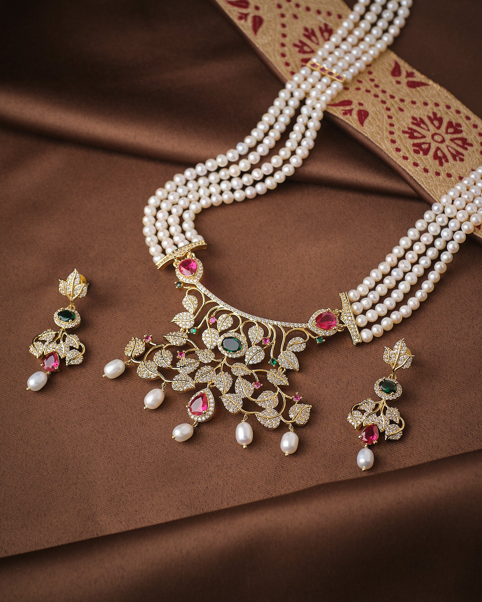 The Noor Floral Supreme Pearl CZ Sita Set by Chandrani Pearls features premium pearls, a gold and gemstone pendant with high-grade CZ stones, and matching earrings, beautifully displayed on brown fabric with a decorative border.