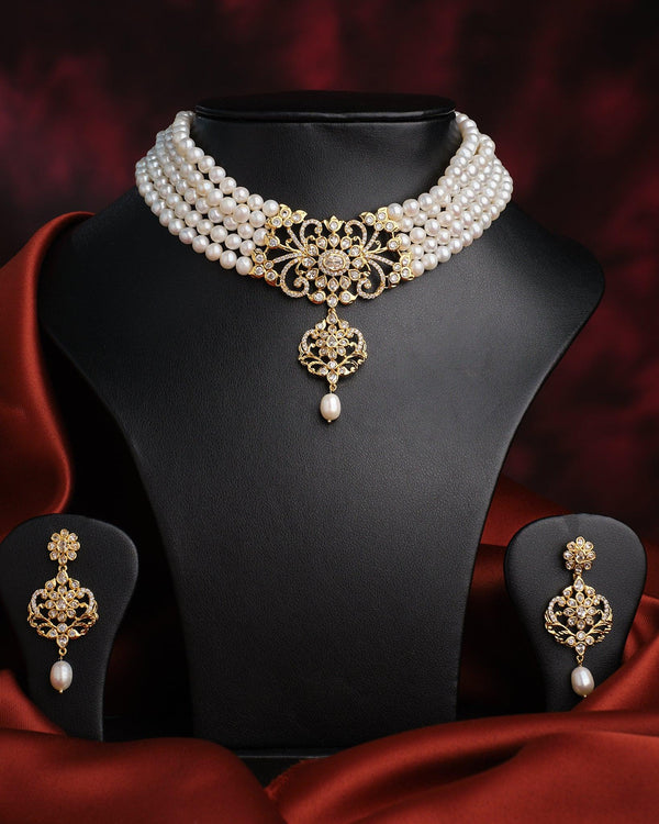 The Modish Victorian Sheen Pearl Necklace Set by Chandrani Pearls features freshwater pearls with a gold clasp, matching pendant, and earrings, beautifully displayed on a black stand against a dark red draped background.