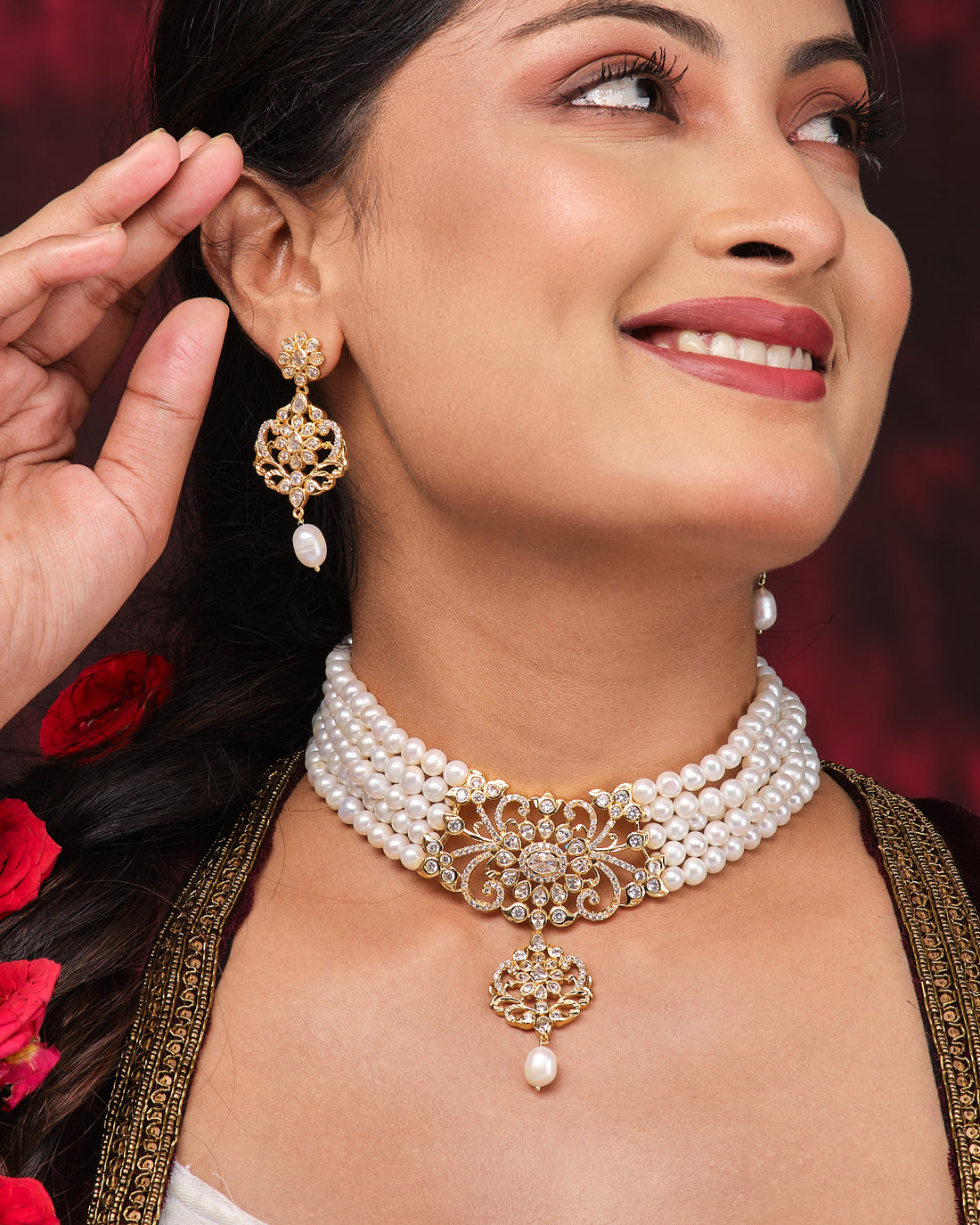The Modish Victorian Sheen Pearl Necklace Set by Chandrani Pearls features freshwater pearls with a gold clasp, matching pendant, and earrings, beautifully displayed on a black stand against a dark red draped background.