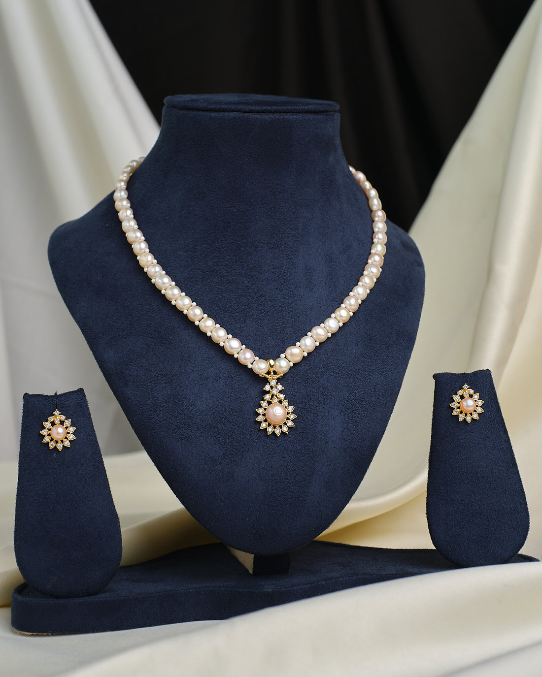 The Romantic Round CZ Necklace Set by Chandrani Pearls features a pearl necklace with a round CZ-accented pendant and matching earrings, elegantly displayed on a navy blue stand against a neutral draped backdrop.