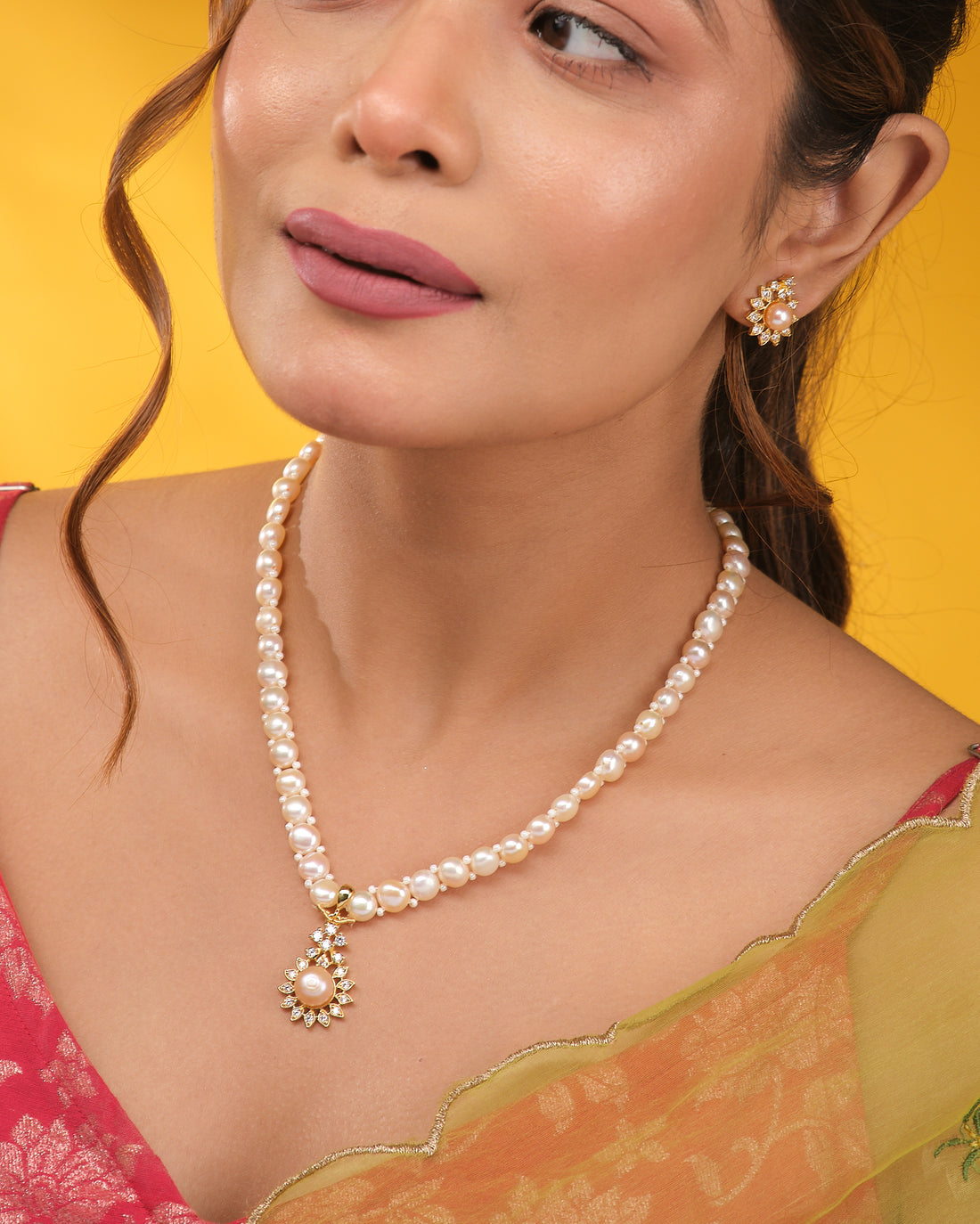 The Romantic Round CZ Necklace Set by Chandrani Pearls features a pearl necklace with a round CZ-accented pendant and matching earrings, elegantly displayed on a navy blue stand against a neutral draped backdrop.