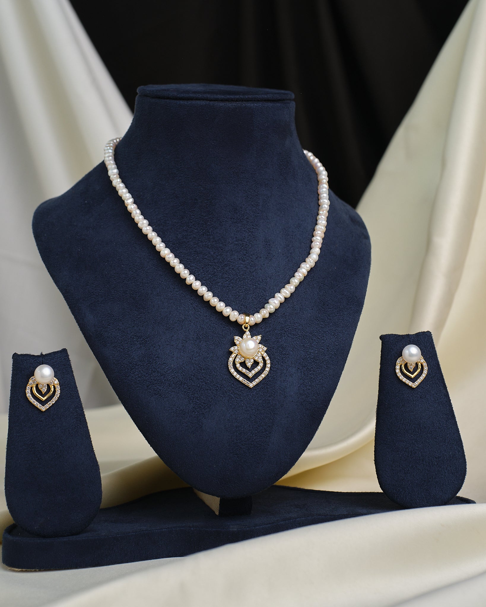 The Chandrani Pearls Elegant Teardrop CZ Set features a pearl necklace with a gold and pearl pendant, displayed on a navy blue stand with matching teardrop earrings, set against draped fabric.