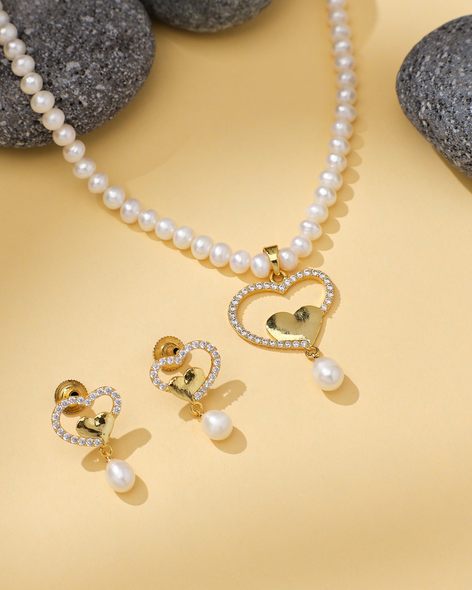 The Queen’s Heart Inspired Pearl CZ Set by Chandrani Pearls features a pearl necklace and matching earrings with gold heart-shaped CZ pendants and hanging pearls, beautifully displayed on a light yellow surface with grey stones.