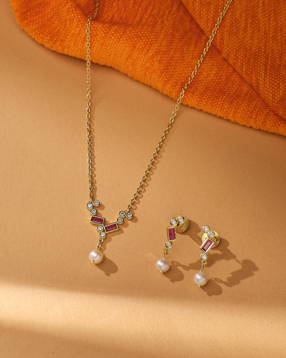 The Pure Elegance Pretty Set by Chandrani Pearls features a gold necklace and matching earrings with geometric designs, red accents, CZ stones, and pearl drops—ideal for formal occasions or gifting—shown on beige with orange textured fabric.
