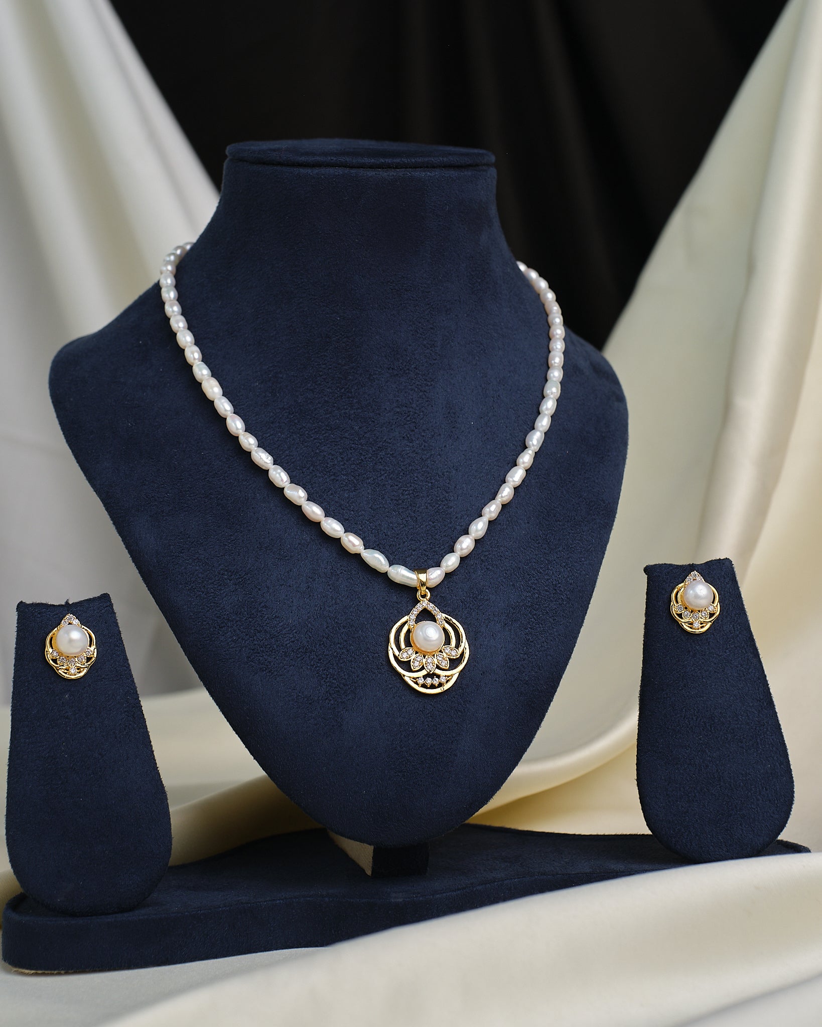 The Elegant Solitaire CZ Necklace Set by Chandrani Pearls is shown on a navy blue stand, featuring a gold and CZ pendant with matching pearl and gold studs on each side.