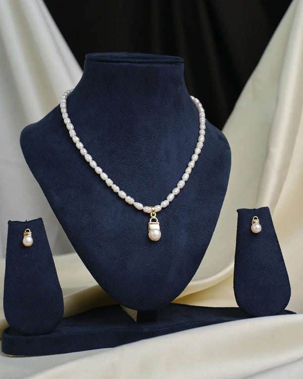 The Round-Cut CZ Tennis Set by Chandrani Pearls, featuring a pearl necklace with a CZ pendant and matching earrings, is displayed on a navy blue jewelry stand against a cream and black fabric background.