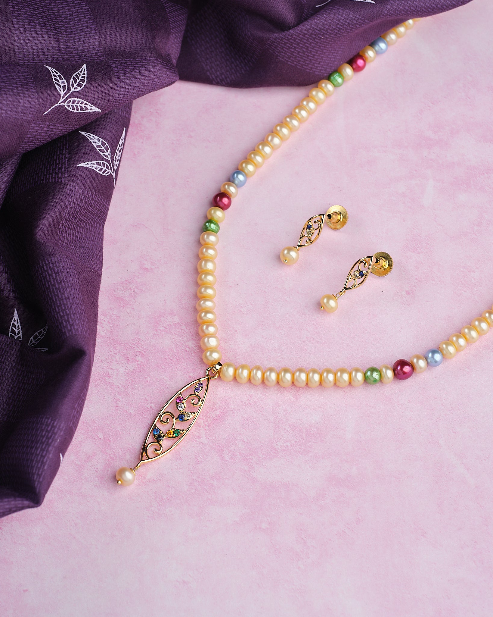Add a pop of color with Chandrani Pearls’ Colorful One-Line Pearl Set with Earrings, featuring vibrant pearls and matching earrings, beautifully displayed on a pink surface beside elegant purple fabric.
