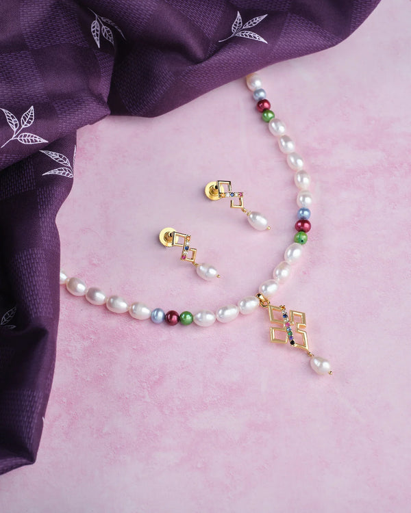 The Regal 1 Line Necklace Set by Chandrani Pearls features glowing pearls, multicolor beads, a gold geometric pendant, and matching gold and pearl earrings, elegantly displayed on a pink surface beside a purple patterned cloth.