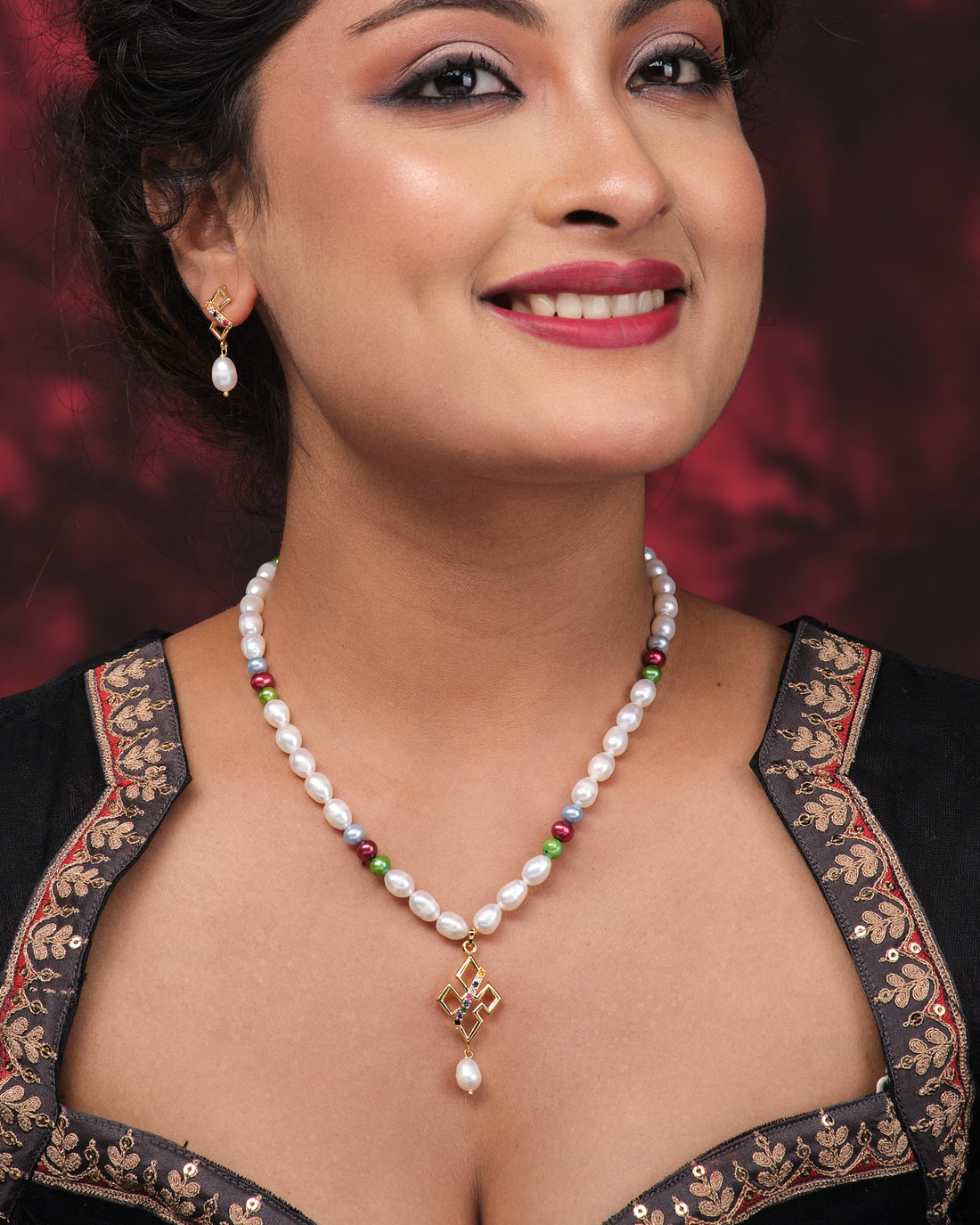 The Regal 1 Line Necklace Set by Chandrani Pearls features glowing pearls, multicolor beads, a gold geometric pendant, and matching gold and pearl earrings, elegantly displayed on a pink surface beside a purple patterned cloth.
