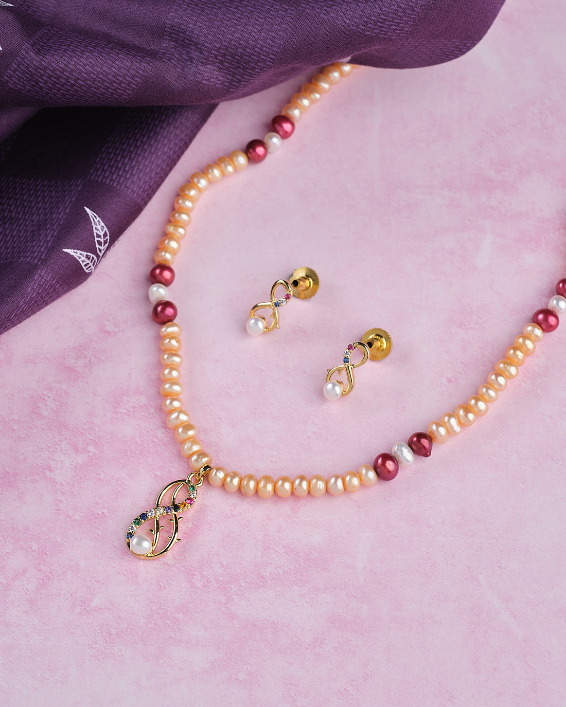 The Elegant Pearl Necklace Set by Chandrani Pearls features a pearl and gold bead necklace with a matching pendant and earrings, displayed on pink with a folded purple cloth, offering subtle design for maximum shine.