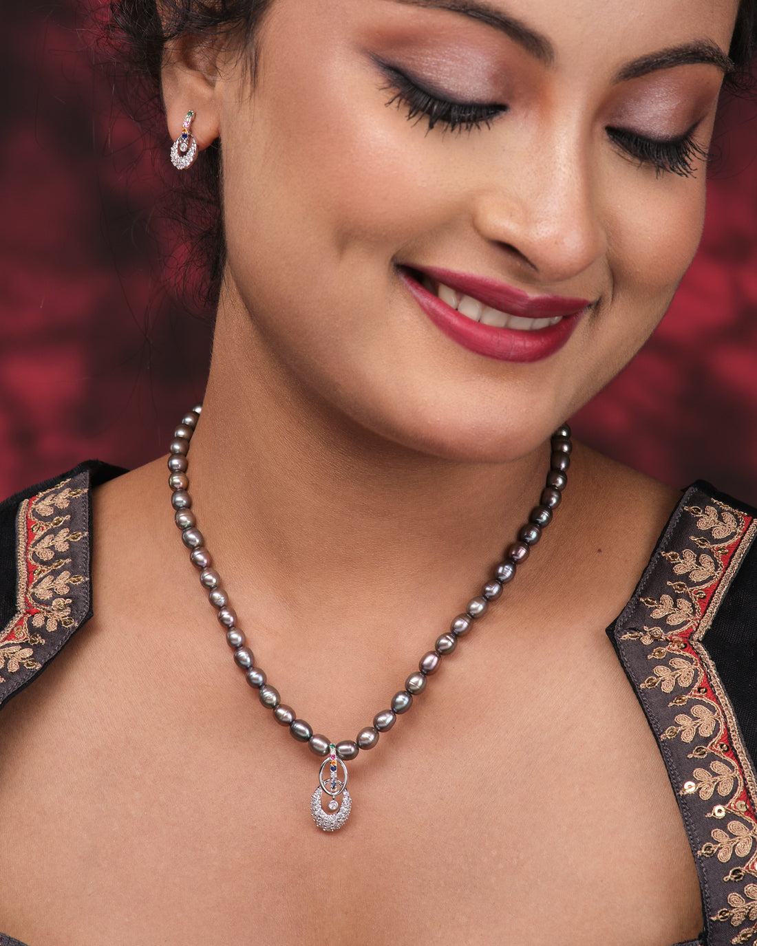 The Chandrani Pearls Vintage Black Stone Simple Pearl Necklace Set, featuring a crystal pendant and matching stud earrings, is displayed on a pink surface beside a folded purple patterned cloth.