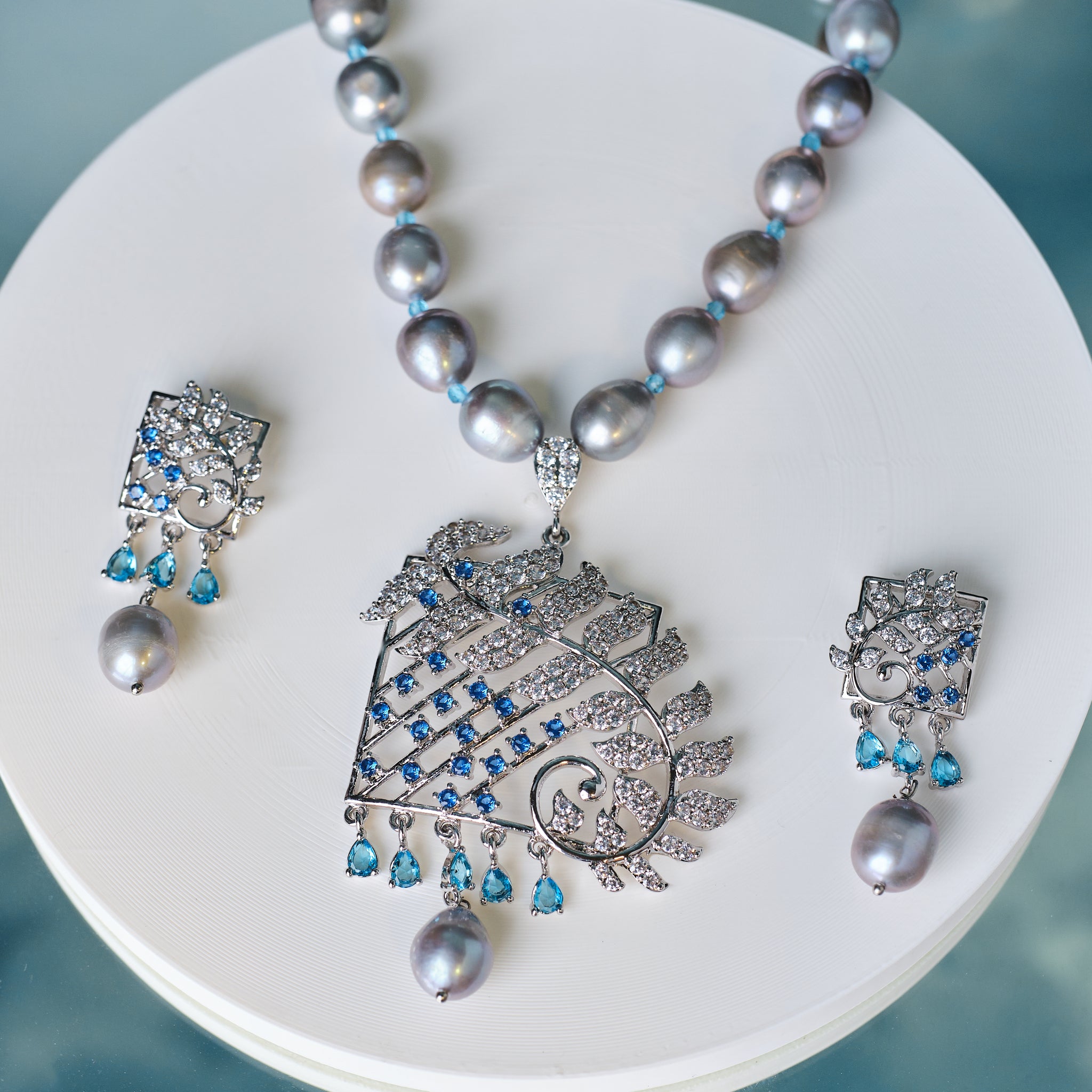 The Chandrani Pearls Dainty Pearl & CZ Jewelry Set includes a gray beaded necklace with an ornate diamond, CZ stones, and blue gemstone pendant, plus matching earrings, displayed on a round white surface.