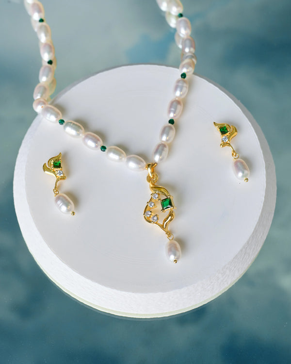 The Statement Pearl & CZ Set by Chandrani Pearls features gold and pearl necklace with matching earrings, green accents, and bold pearls—displayed on a white platform against a cloudy blue background, perfect for standout occasions.