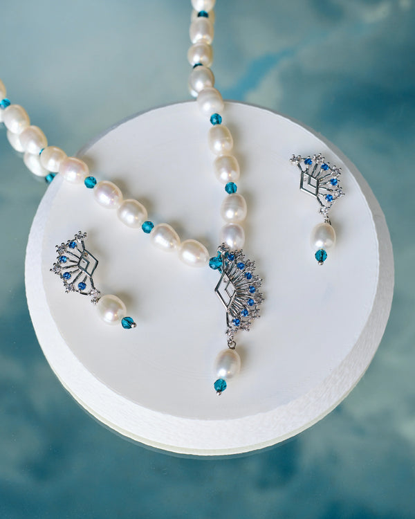 The Chandrani Pearls Evening Pearl & CZ Necklace Set, featuring a pearl and blue gemstone necklace with matching earrings, is elegantly displayed on a white platform against a blue background—ideal for evening occasions.