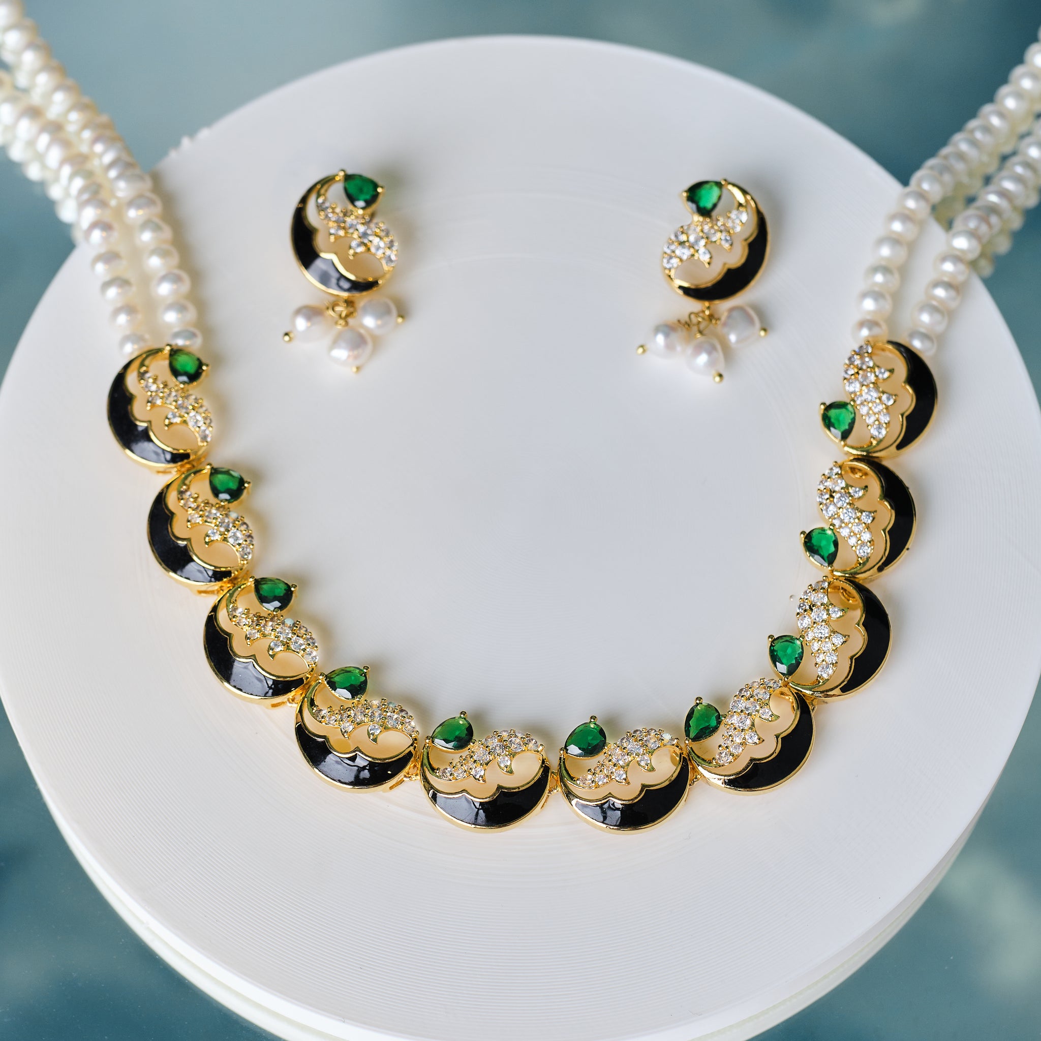 The Gorgeous Pearl & CZ Stone Necklace Set by Chandrani Pearls features matching earrings with green CZ stones and white pearls, displayed on a white round stand against a blue background.