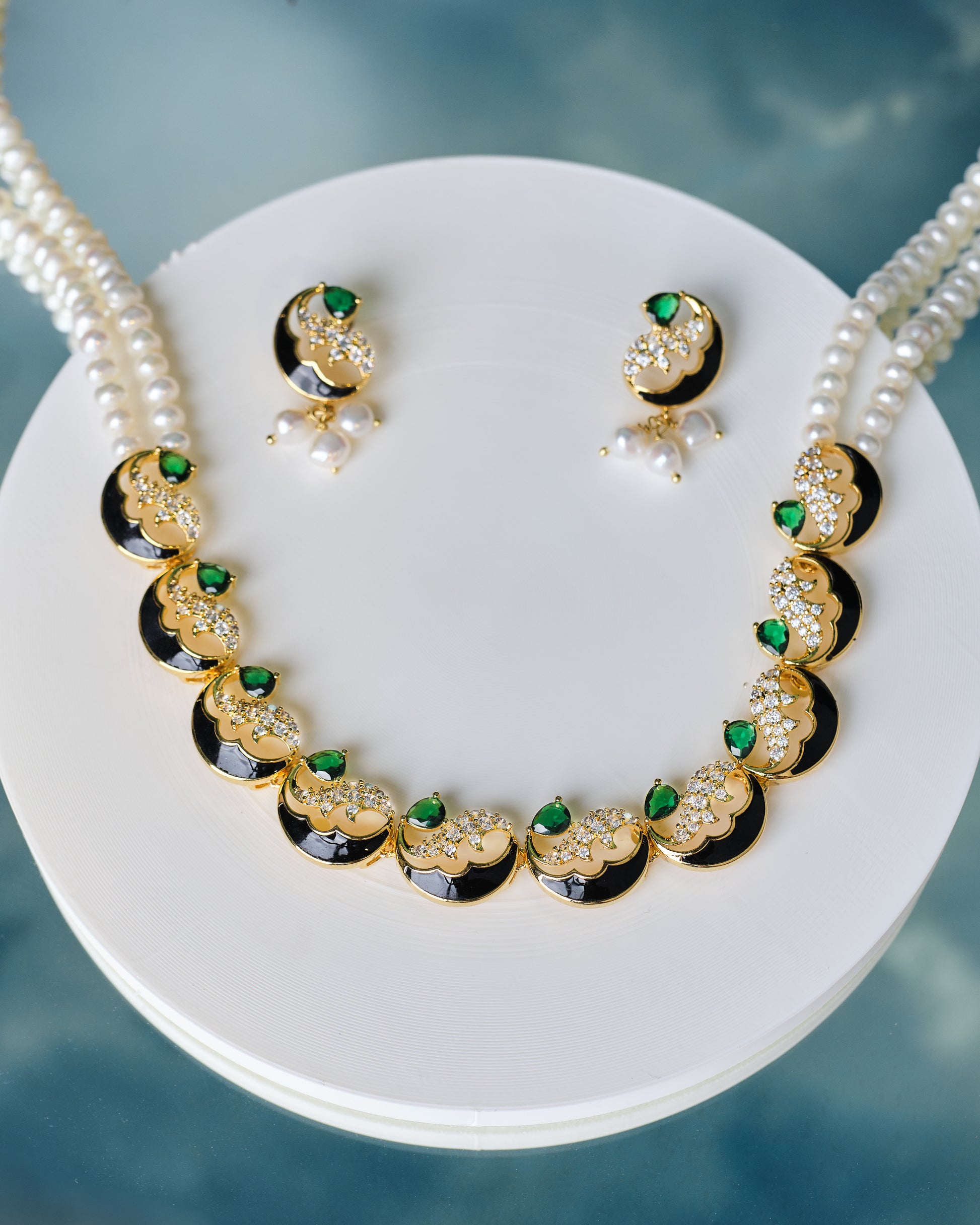 The Gorgeous Pearl & CZ Stone Necklace Set by Chandrani Pearls features matching earrings with green CZ stones and white pearls, displayed on a white round stand against a blue background.