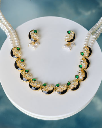 The Gorgeous Pearl & CZ Stone Necklace Set by Chandrani Pearls features matching earrings with green CZ stones and white pearls, displayed on a white round stand against a blue background.