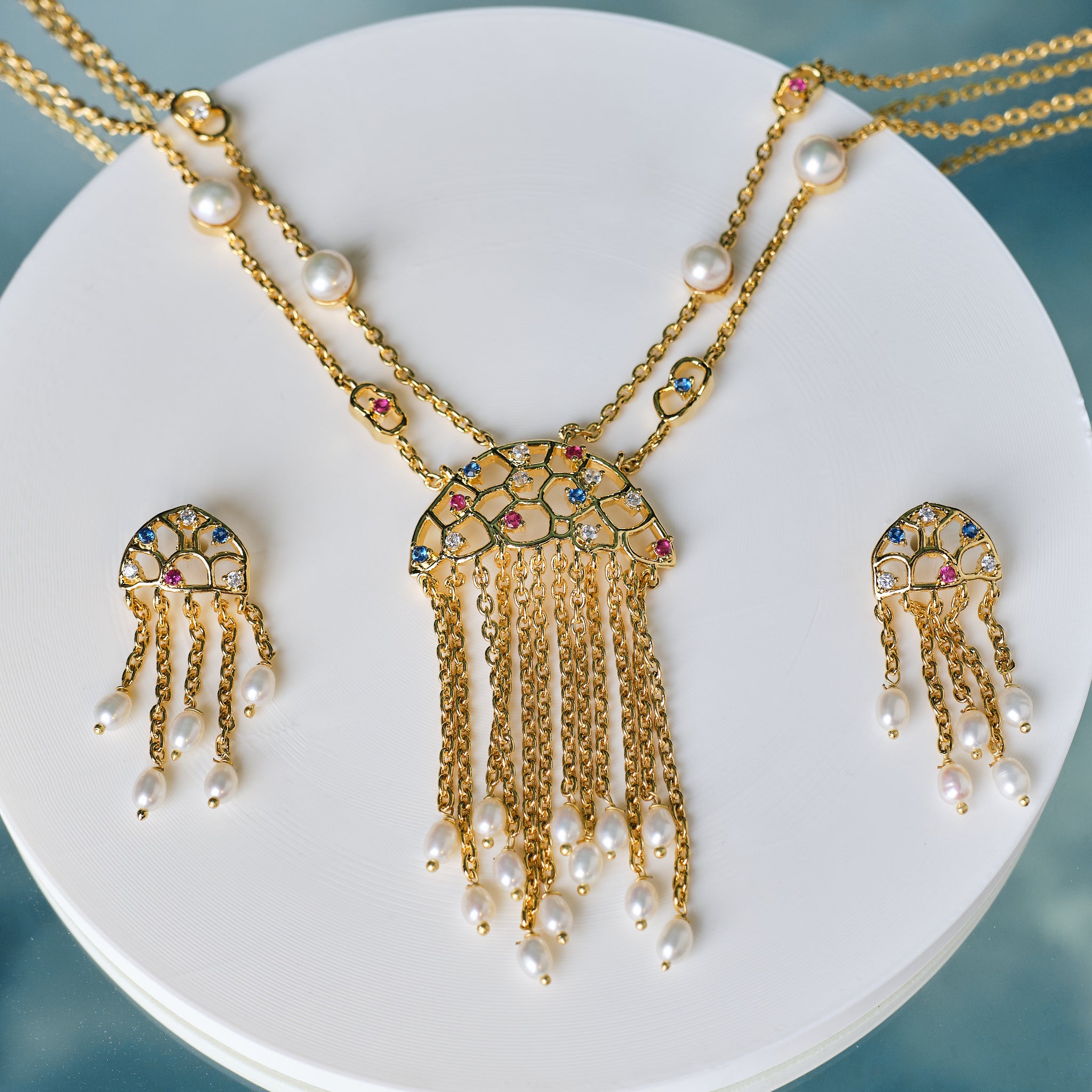 The Trendy Handcrafted Chain & CZ Necklace Set by Chandrani Pearls features colorful gemstones, CZ stones, and pearls in a multi-chain design with matching earrings, elegantly displayed on a white circular stand.