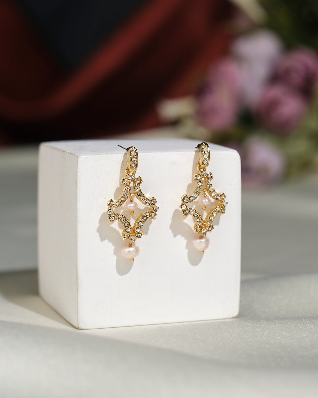 Delicate Pretty CZ Earrings by Chandrani Pearls India, featuring a classic design with rhinestones and pearl drops, are displayed on a white cube against a blurred floral background.