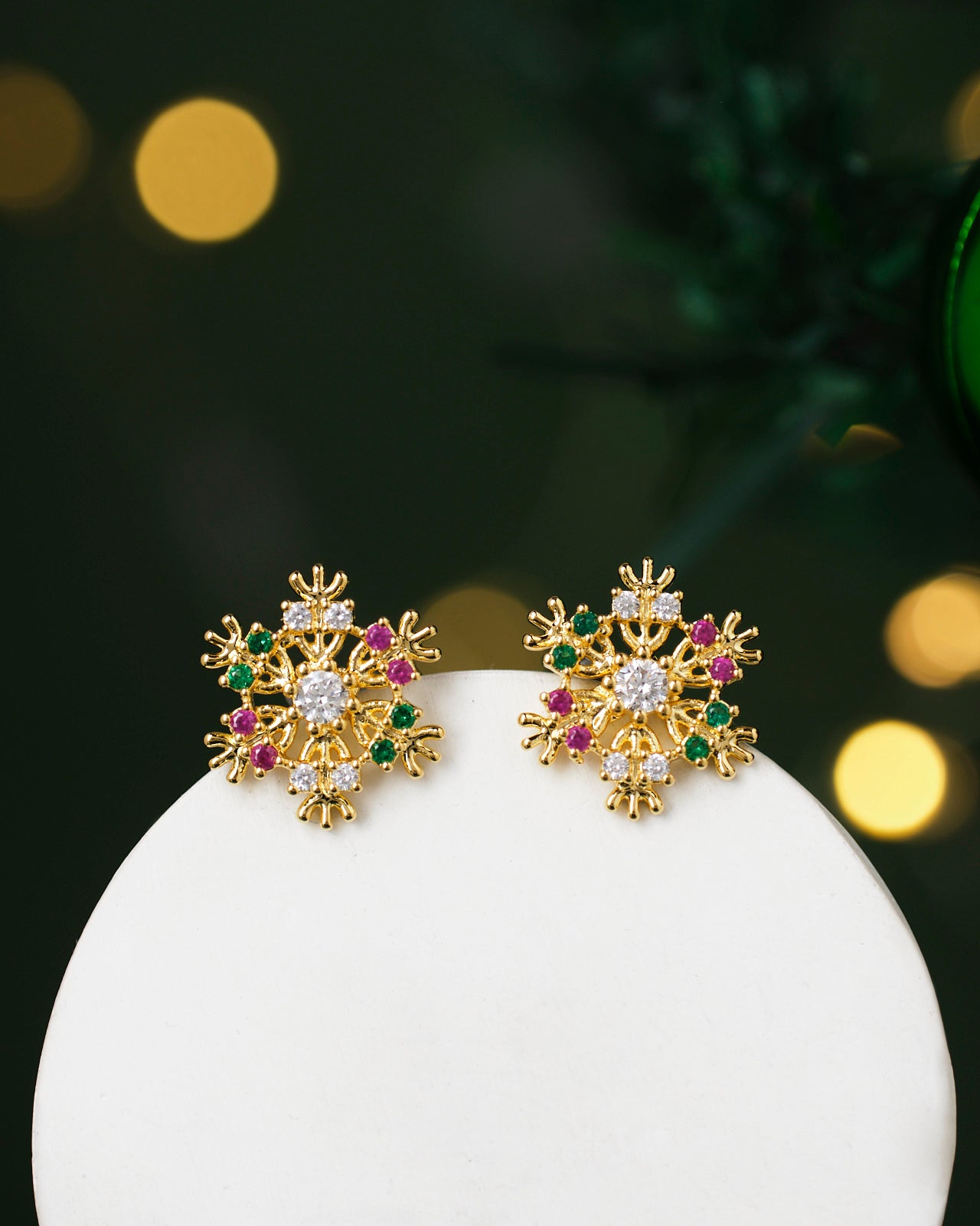 Cuteness Amplified Earrings from Chandrani Pearls: gold, snowflake-shaped with red, green, and clear gemstones on a white surface with blurred background lights.