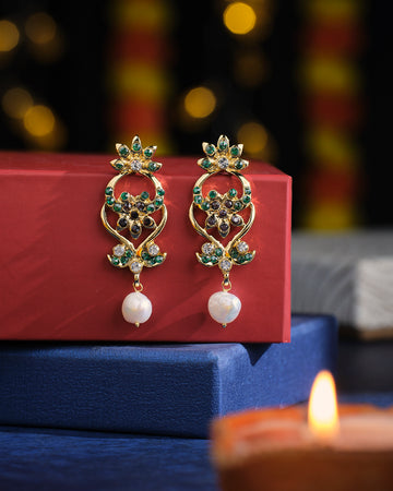 Holiness Shine Pearl Earrings by Chandrani Pearls, featuring gold and emerald designs with pearl drops, displayed on red and blue boxes. A lit candle sits in the foreground.