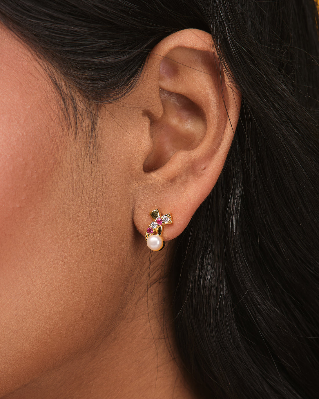 The Elegant Bloom CZ Pearl Earrings by Chandrani Pearls feature gold cross flower studs with pink stones, CZ accents, and a round white pearl on each, displayed on a neutral surface.