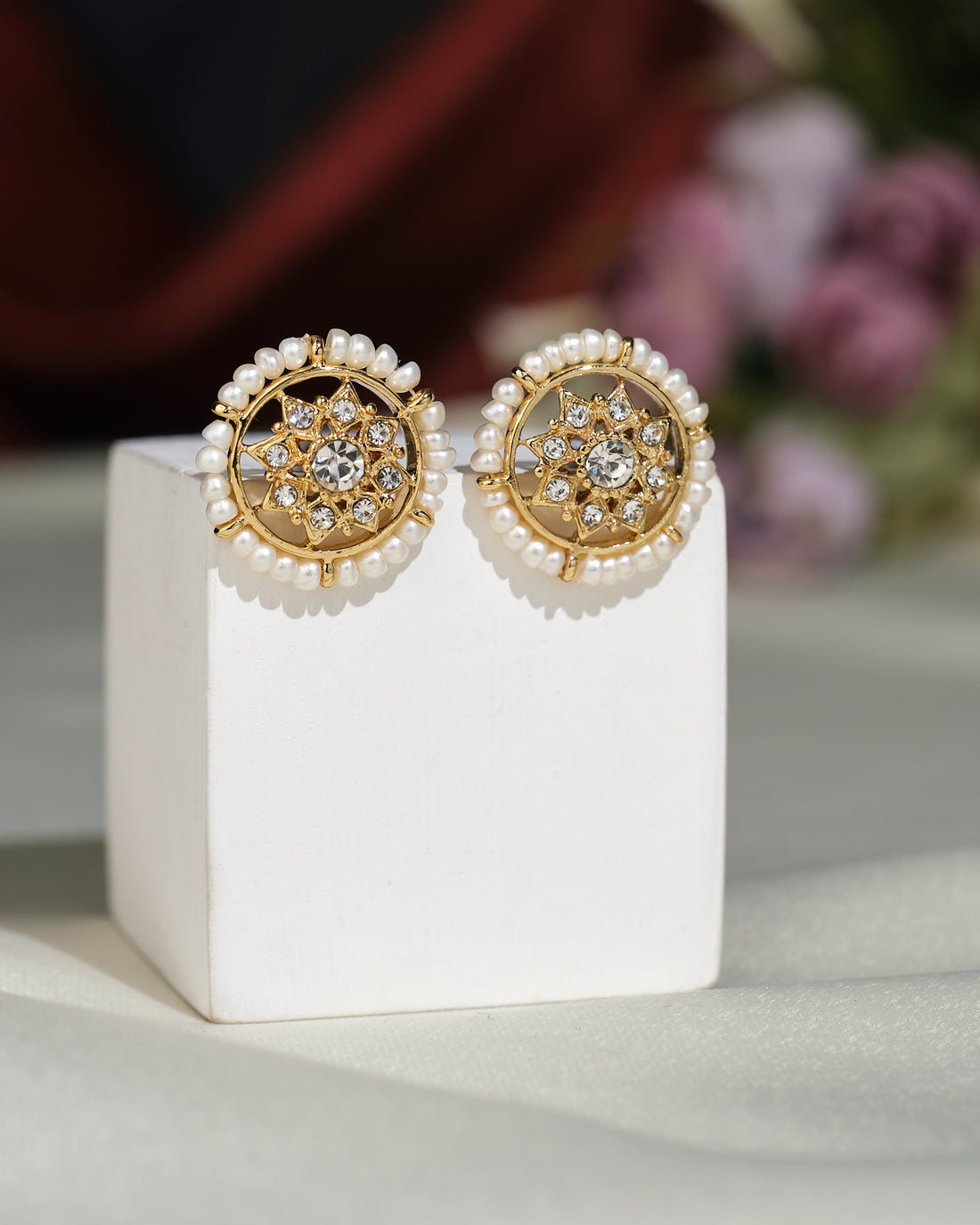 Traditional Beautiful CZ Earrings by Chandrani Pearls India, featuring round gold pearls with crystal accents, are elegantly displayed on a white stand against a blurred floral background.
