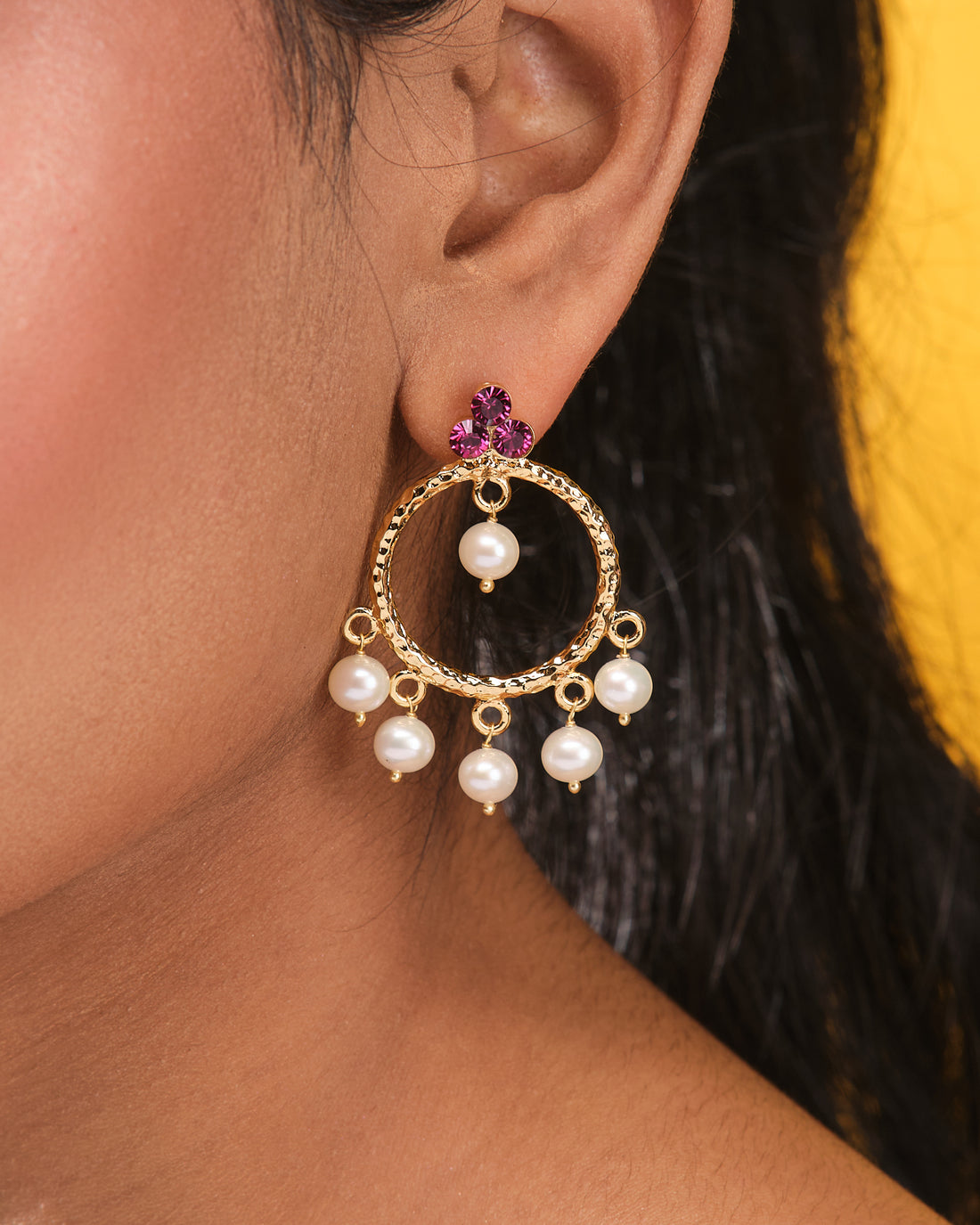 Chandrani Pearls Round CZ Studs with Pearl Dangle Modern Glam—gold hoop earrings featuring purple flower-shaped CZ studs and pearl drops, displayed on a beige surface beside smooth black stones for classic elegance.
