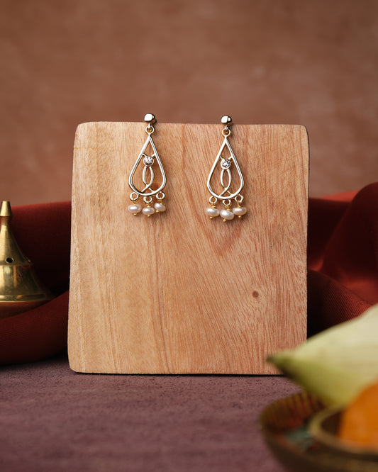 The Trendy Pearl Earrings by Chandrani Pearls India, featuring a teardrop design with pearl accents, are elegantly displayed on a wooden stand against a brown background adorned with decorative objects.