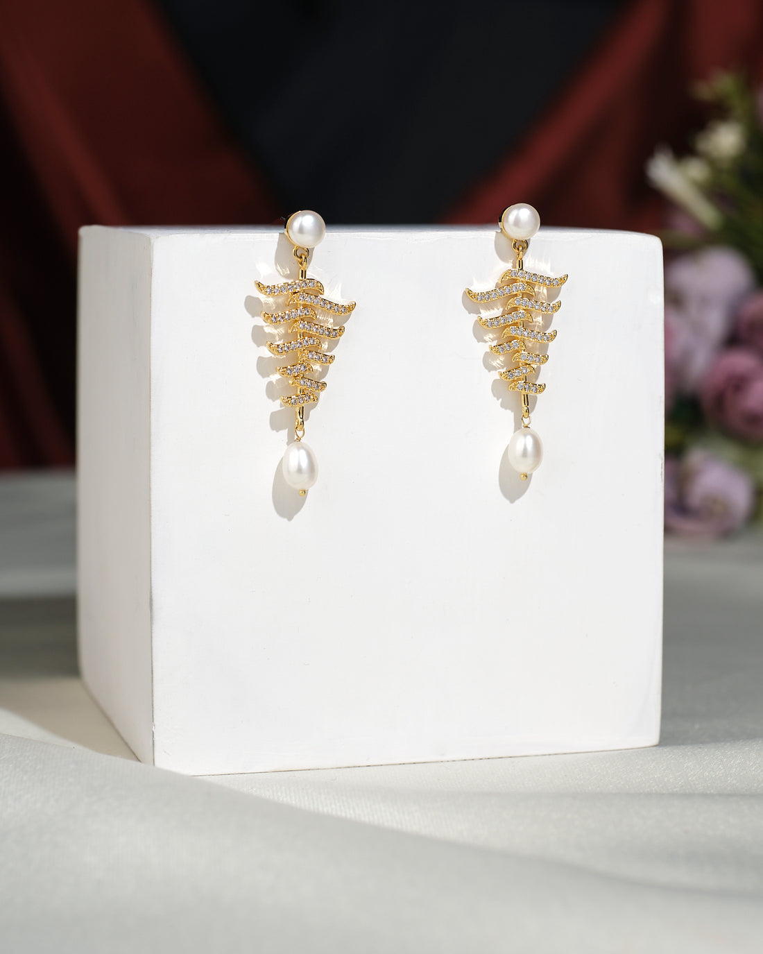 Vintage Hang Earrings by Chandrani Pearls India feature a gold zigzag design with pearl accents, showcased on a white cube for a touch of elegance against a softly blurred floral background.