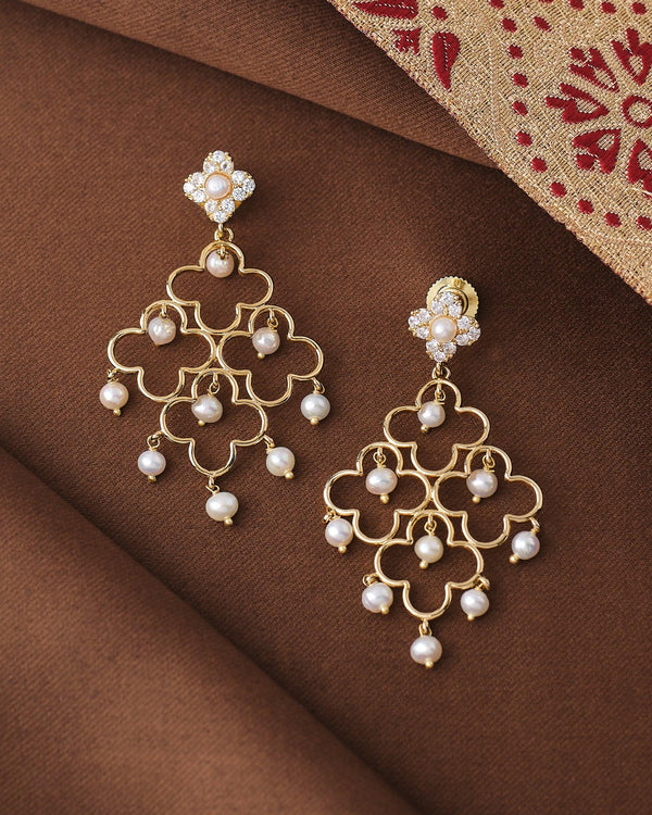 The Noor Radiance Pearl Drop Earrings by Chandrani Pearls feature a detailed flower design with pearl drops, displayed on brown fabric beside a patterned textile.