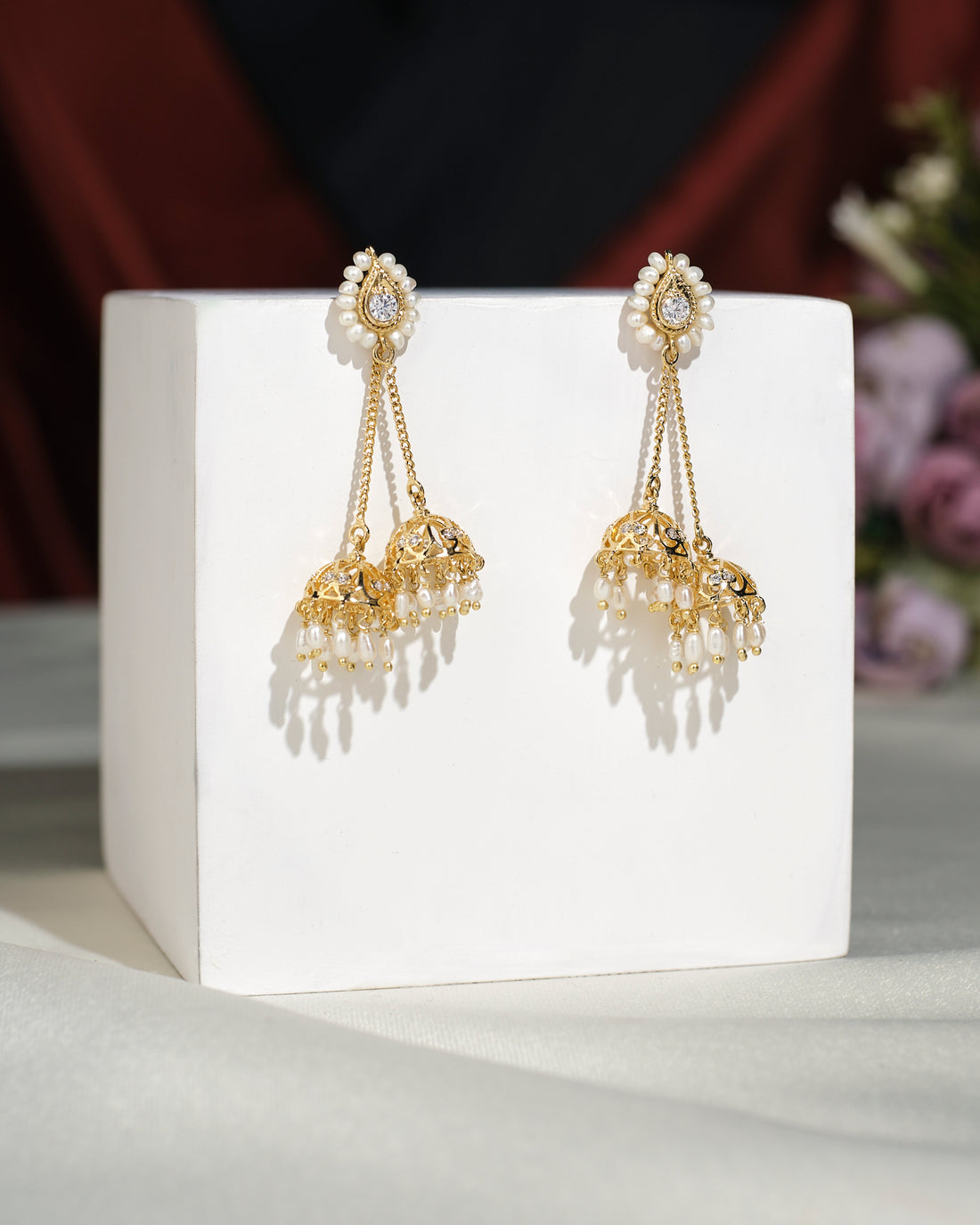 Beautiful Double Jhumka earrings by Chandrani Pearls India, with gold finish and pearl accents in a classic design, displayed on a white cube against a blurred floral background.