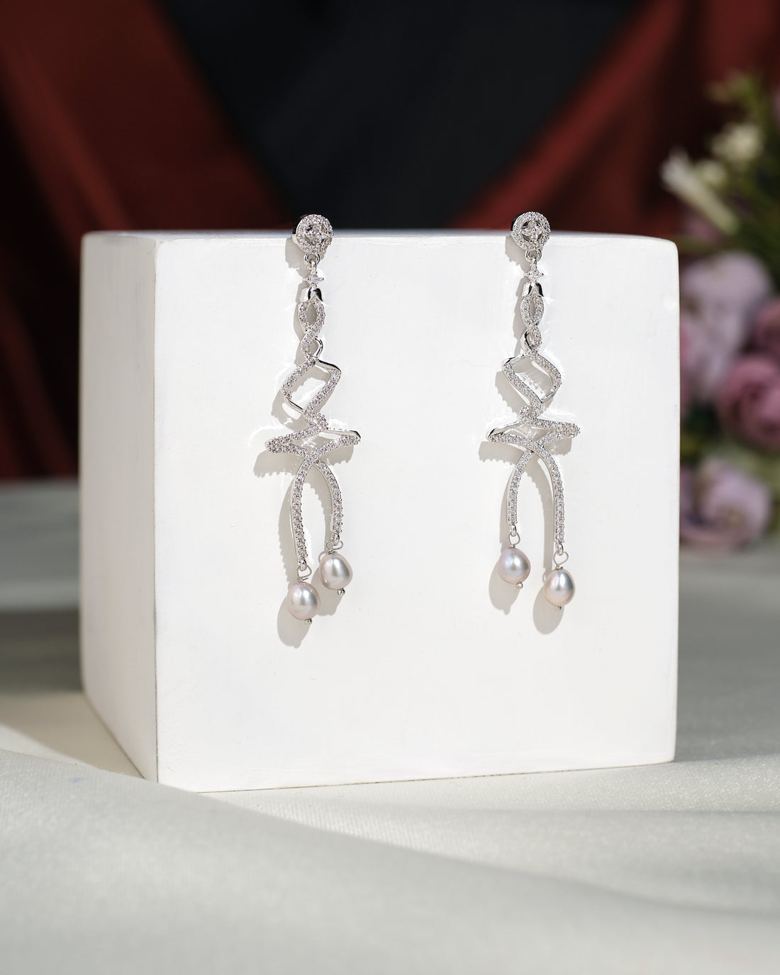 The Trendy CZ Hang Earrings by Chandrani Pearls India feature elegant ribbon-inspired silver dangles with pearl drops, showcased on a white cube against a soft floral backdrop.