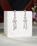 The Trendy CZ Hang Earrings by Chandrani Pearls India feature elegant ribbon-inspired silver dangles with pearl drops, showcased on a white cube against a soft floral backdrop.