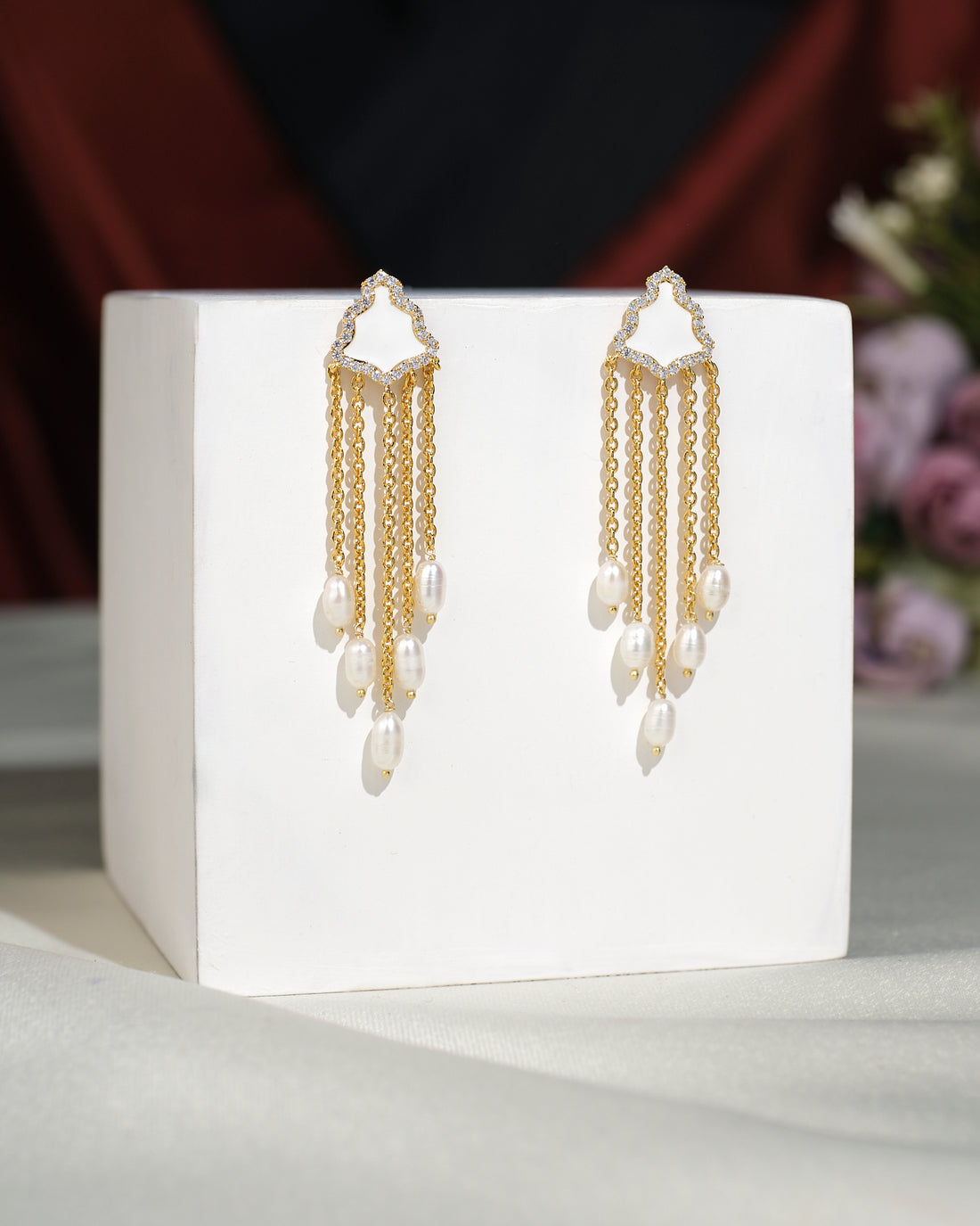 The Stylish Hang Earrings by Chandrani Pearls India, featuring gold and pearls, are displayed on a white cube with a blurred floral background.