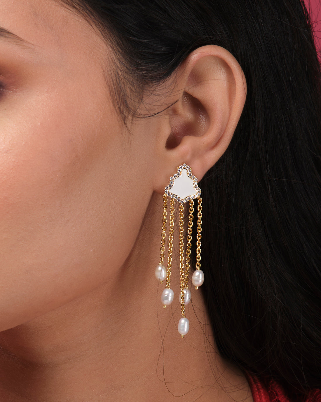 The Stylish Hang Earrings by Chandrani Pearls India, featuring gold and pearls, are displayed on a white cube with a blurred floral background.