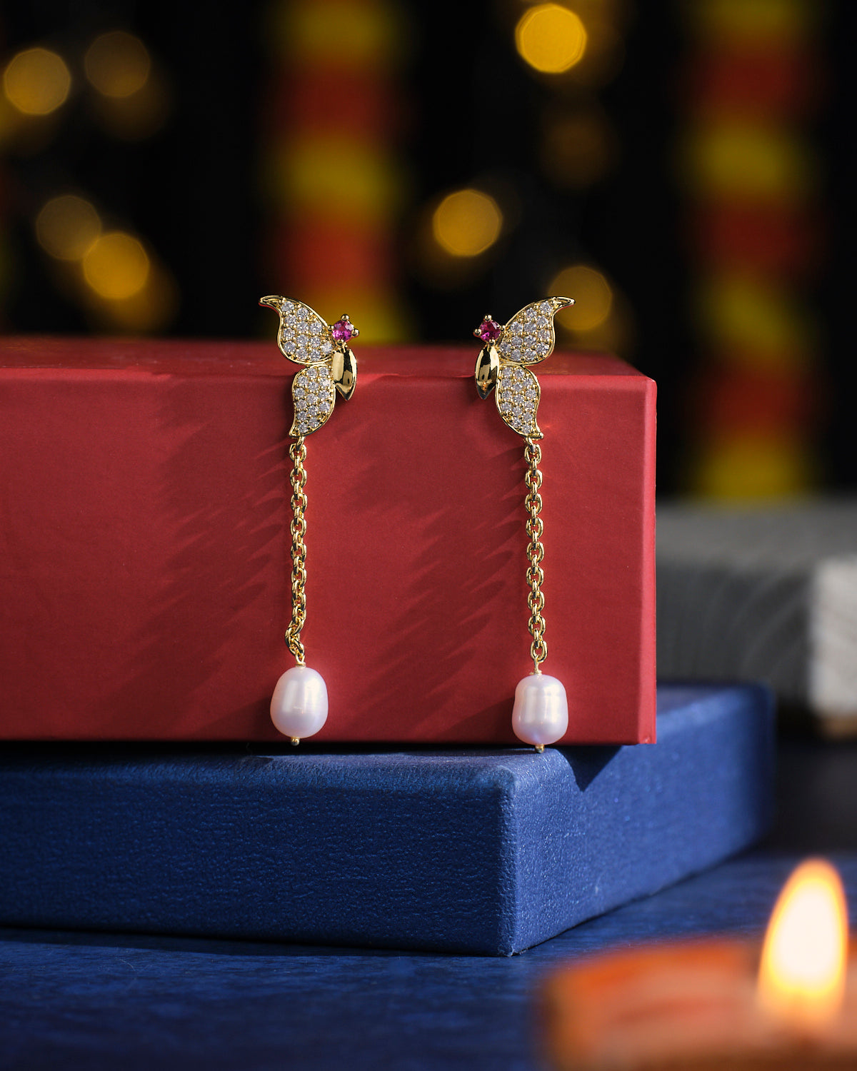 The **Charismatic Half Butterfly Pearl Earrings** by **Chandrani Pearls** are showcased on stacked red and blue boxes, with blurred colorful lights creating a vibrant backdrop.